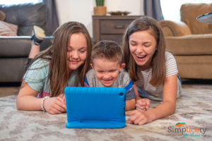 Three children laying on a floor watching a tablet with their hair touching